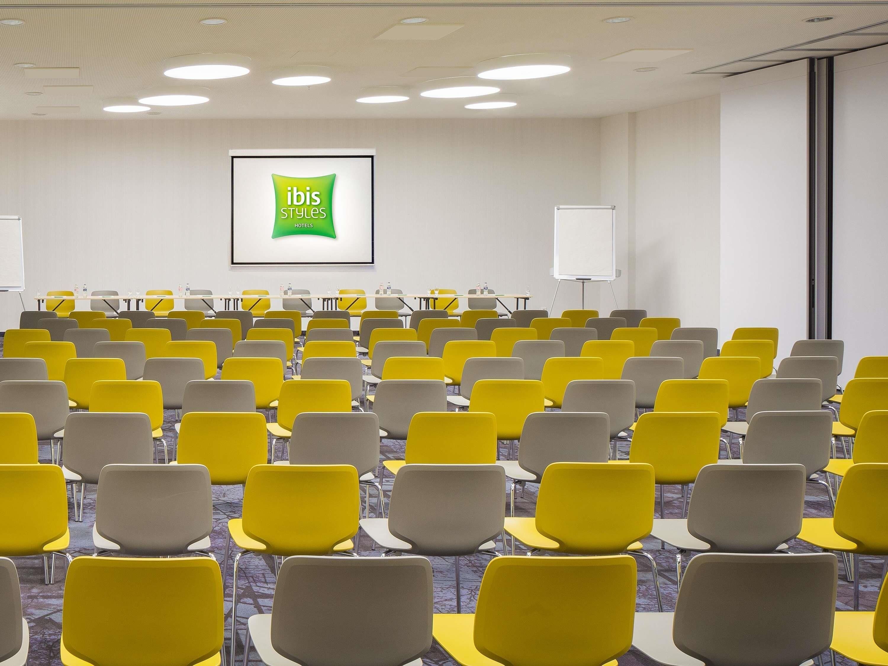 A conference room with rows of yellow chairs, a whiteboard, and a screen displaying 'ibis Styles Hotels'.