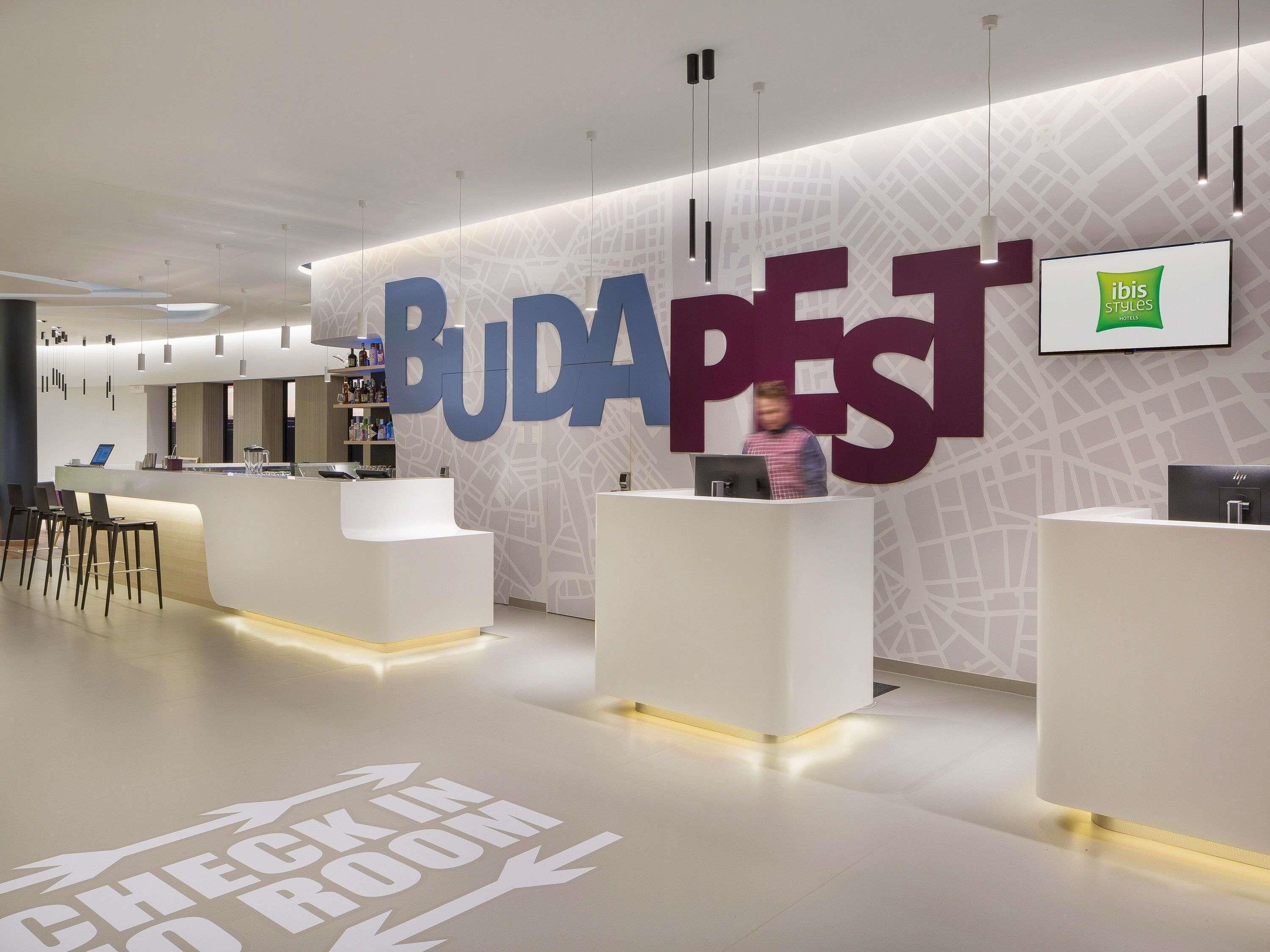 Una zona de recepción moderna con un cartel grande que dice 'BUDAPEST' y una señalización en el suelo que indica 'CHECK IN ROOM'.