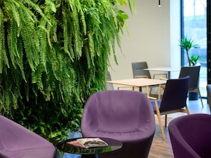 Un espacio interior moderno con un gran muro cubierto de plantas verdes, asientos morados y una pequeña mesa con revistas.