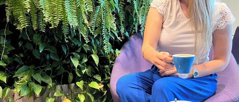 A person sitting on a purple couch with a dog beside them, holding a blue cup, in a room with a large fern wall and a round glass table.