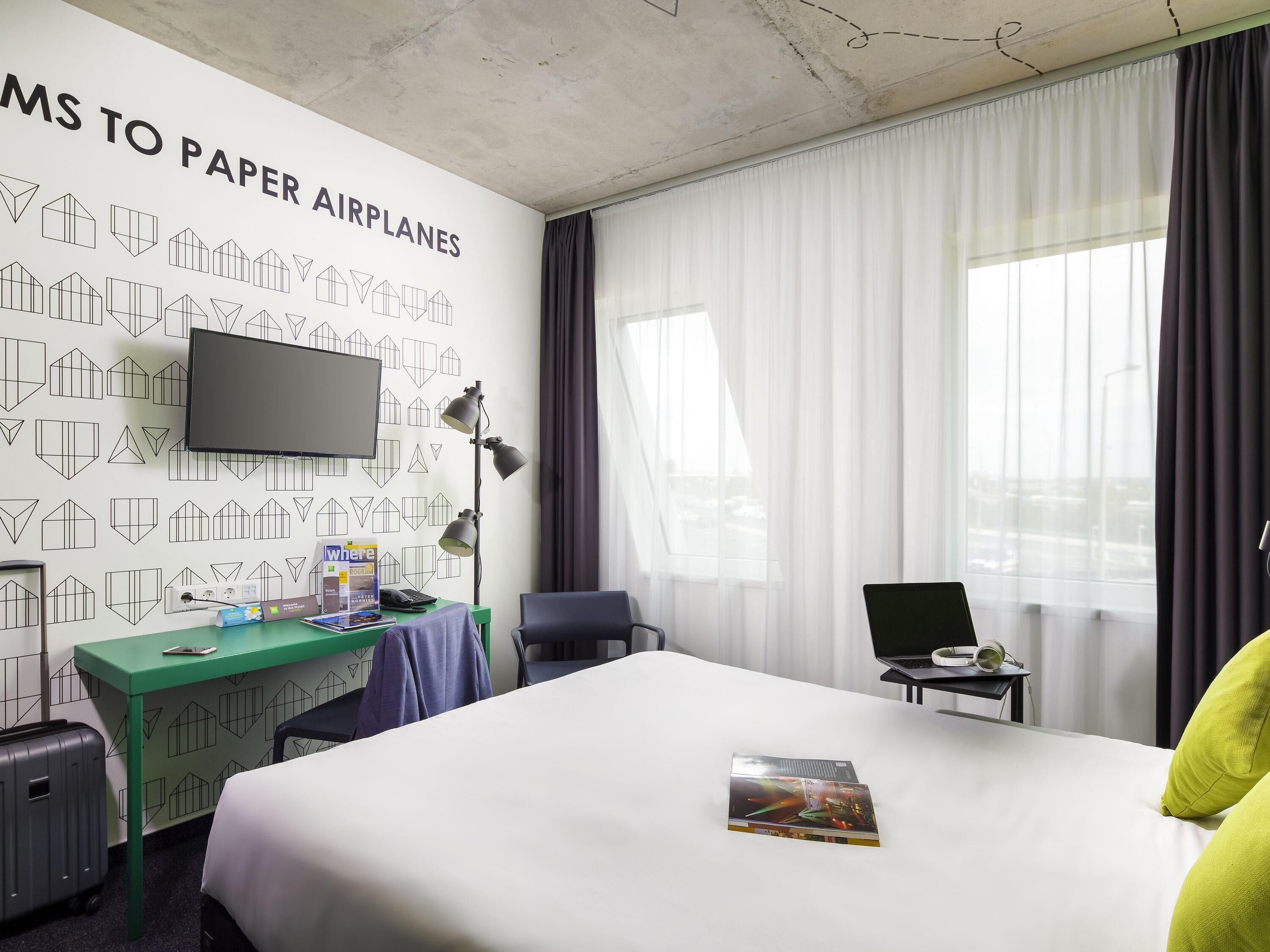 A hotel room with a bed, a desk with a computer, a TV, and a window with curtains.