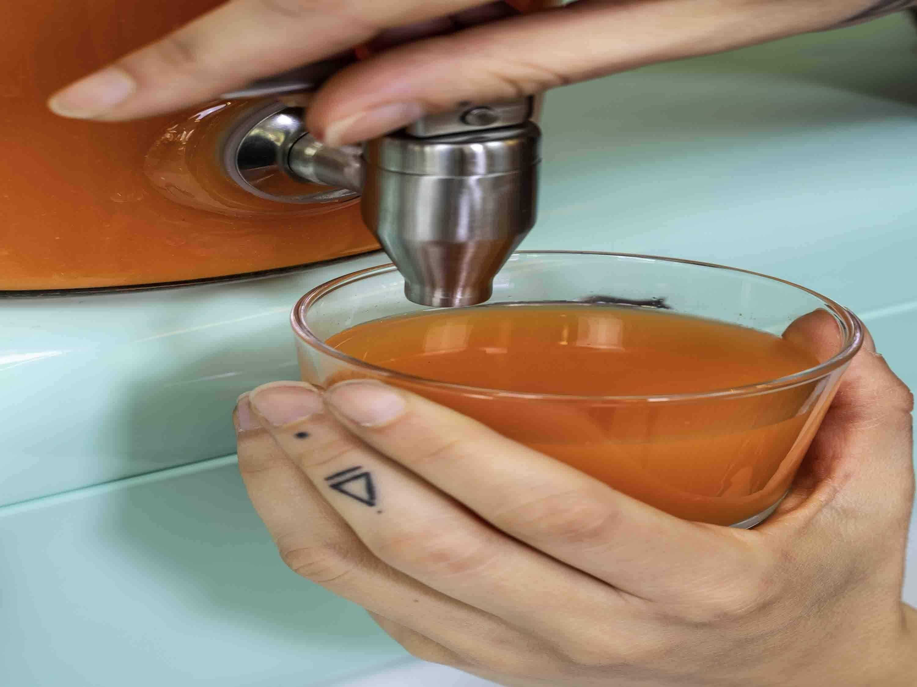 A hand holding a glass with a liquid being poured from a metallic dispenser.