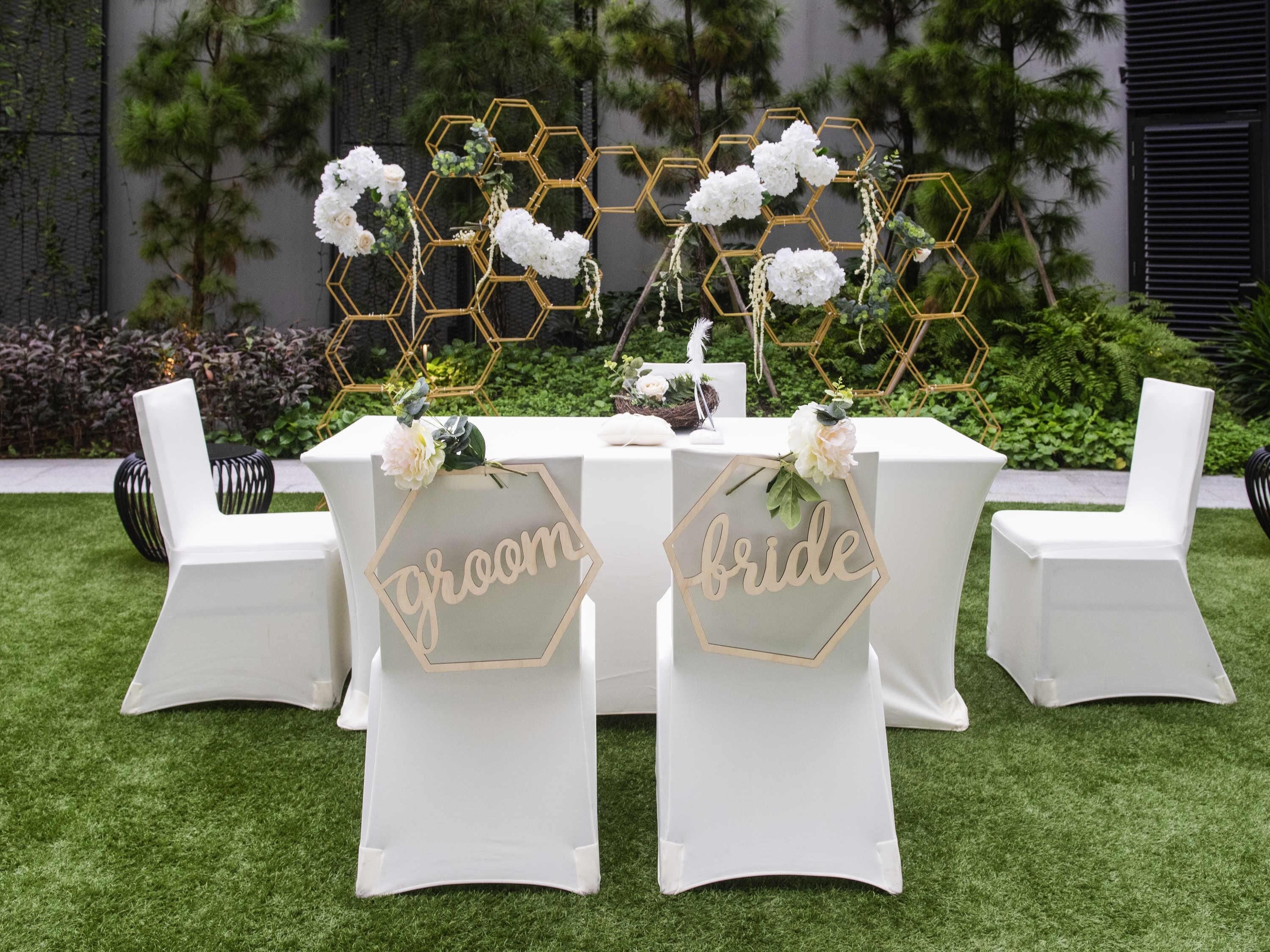 A wedding reception table with hexagonal gold decorations, white floral arrangements, and chairs labeled 'groom' and 'bride.'