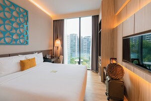 A hotel room with a large bed, a desk, a chair, a lamp, a suitcase, and a large window with a view of buildings.