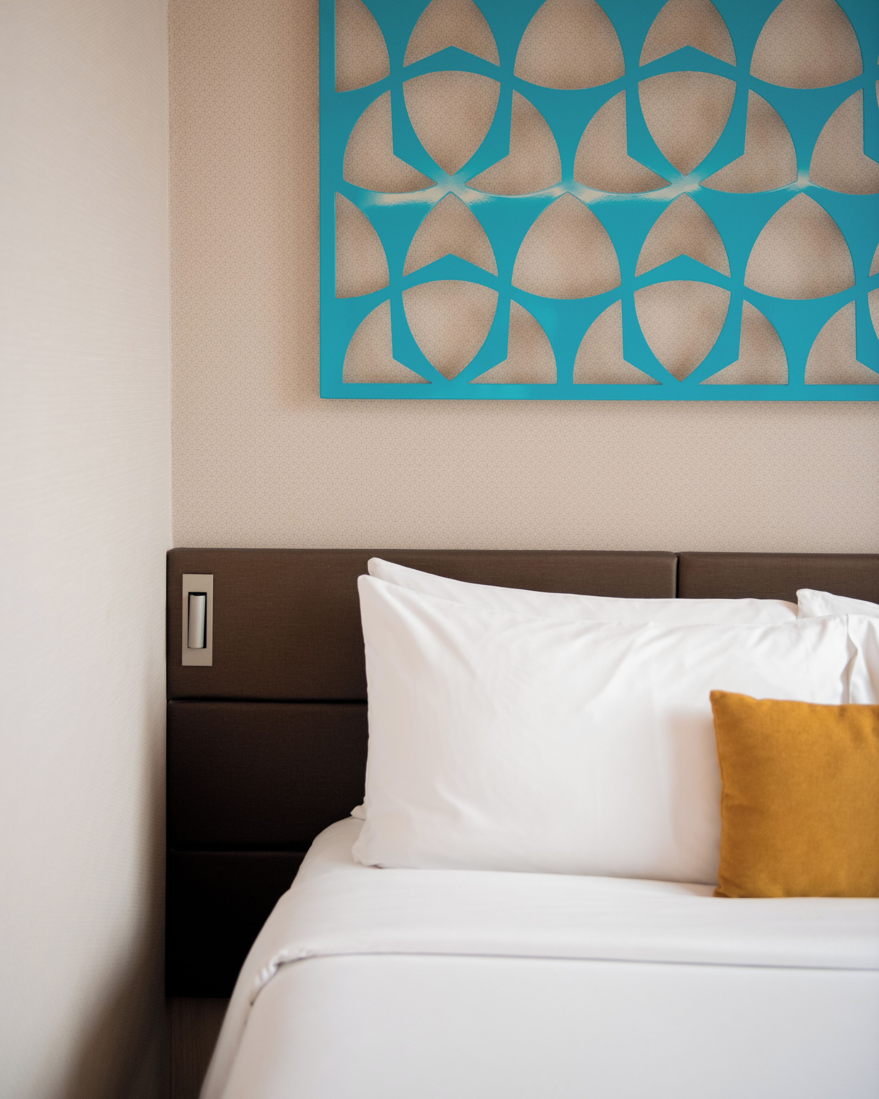 Un letto rifatto con cuscini bianchi e color senape, una testiera marrone e un pannello decorativo sulla parete sovrastante.