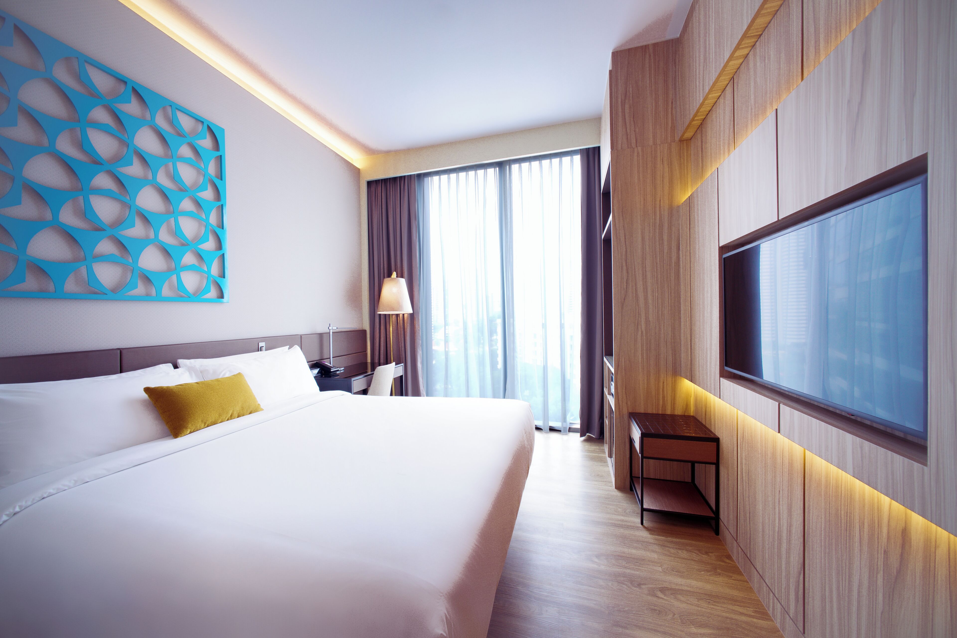 A modern hotel room with a large bed, a desk, a TV, and a view of the city through large windows.