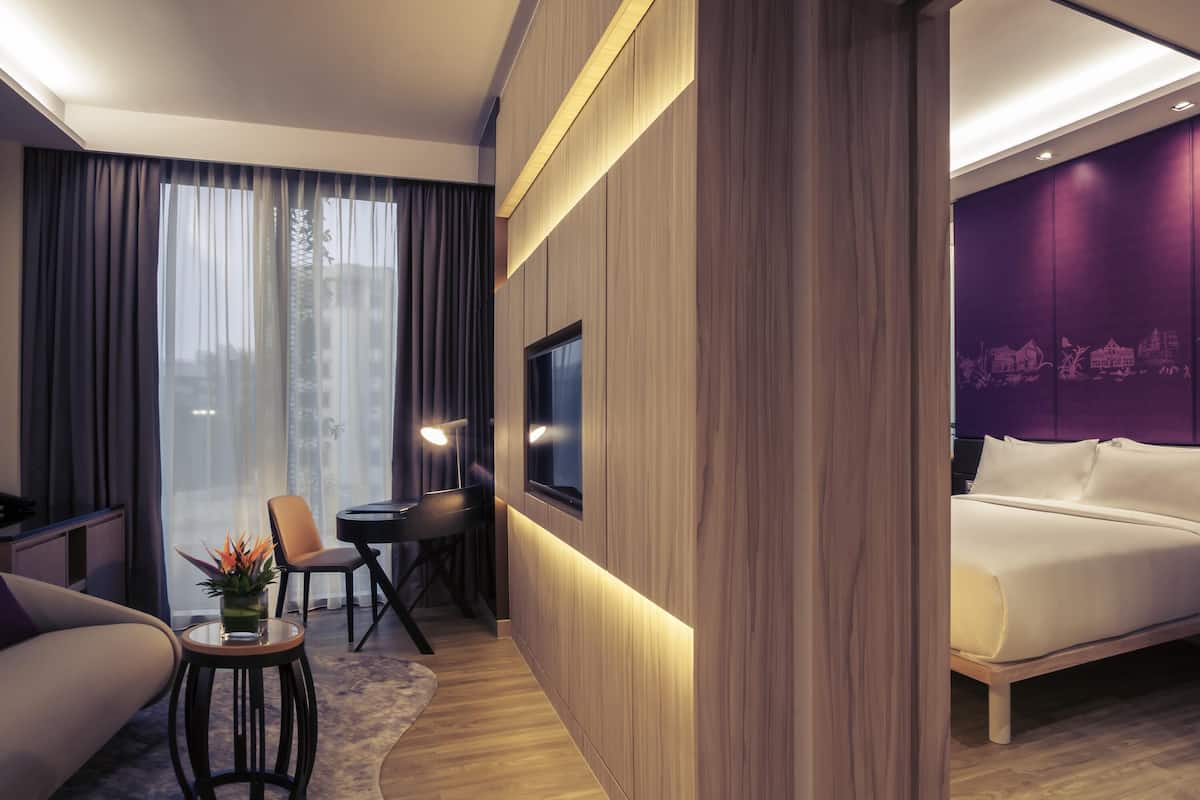 A modern hotel room with a bed, a desk with a chair, a small table with a plant, and a large window with curtains.