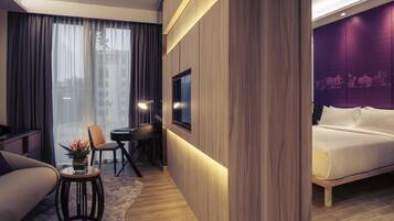 A modern hotel room with a bed, a desk with a chair, a small table with a plant, and a large window with curtains.
