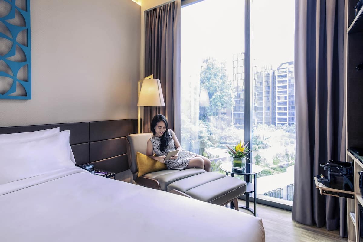 Quarto de hotel com uma cama grande, um sofá, uma mesinha de centro com abajur e vista para edifícios pela janela.