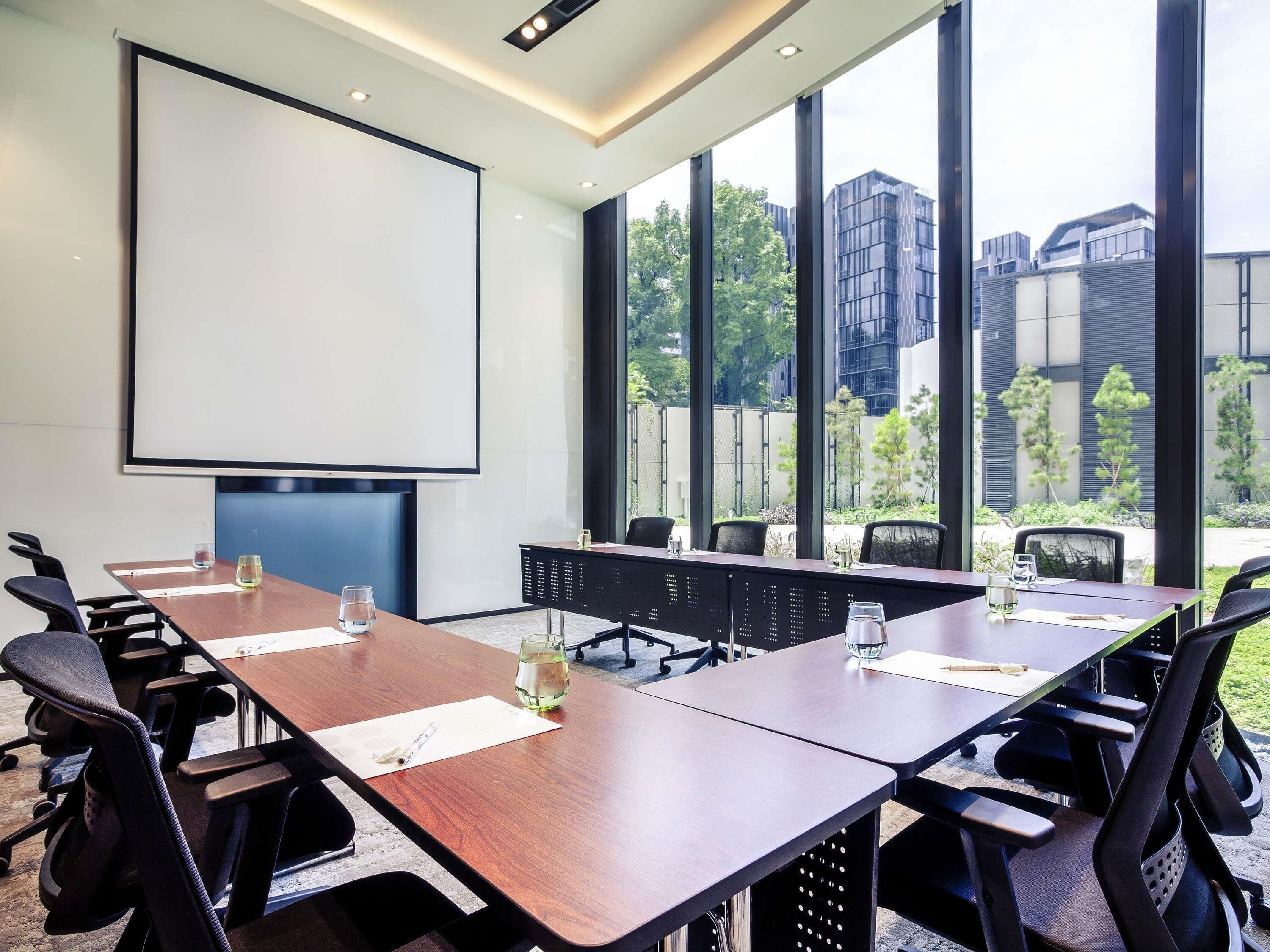 Ein Konferenzraum mit großem Tisch, Stühlen, einer Projektionsleinwand und großen Fenstern mit Blick auf Gebäude und Bäume.