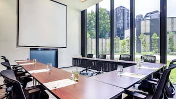 Ein Konferenzraum mit großem Tisch, Stühlen, einer Projektionsleinwand und großen Fenstern mit Blick auf Gebäude und Bäume.