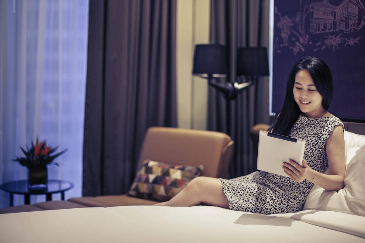 Uma mulher sentada em uma cama num quarto de hotel, lendo um livro.
