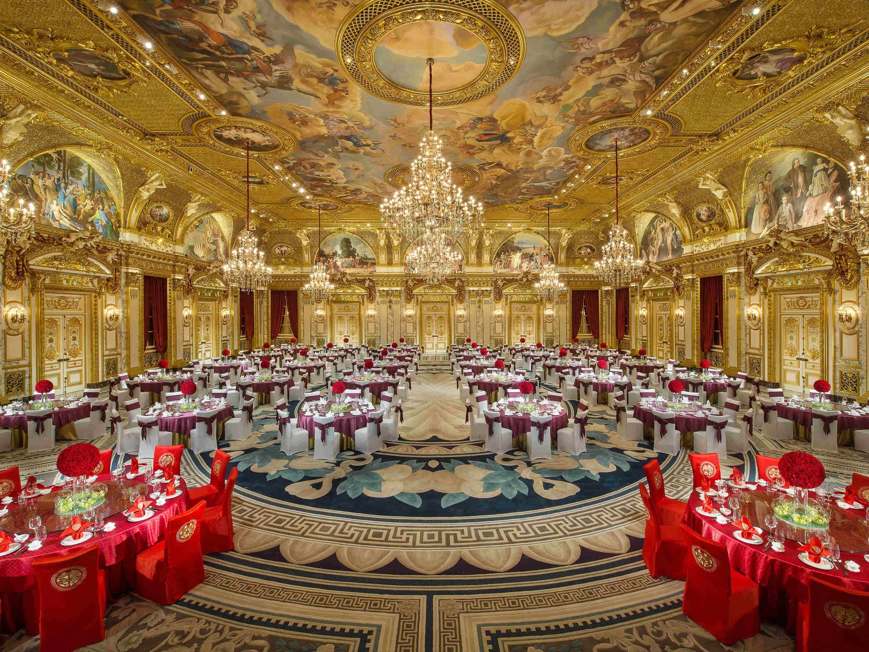 Un gran salón de banquetes con frescos ornamentales en el techo, candelabros y mesas redondas dispuestas para un evento formal.