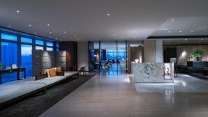 Un hall d’hôtel moderne doté d’un long comptoir d’accueil, d’un espace salon avec des canapés et de grandes fenêtres offrant une vue sur la ville.
