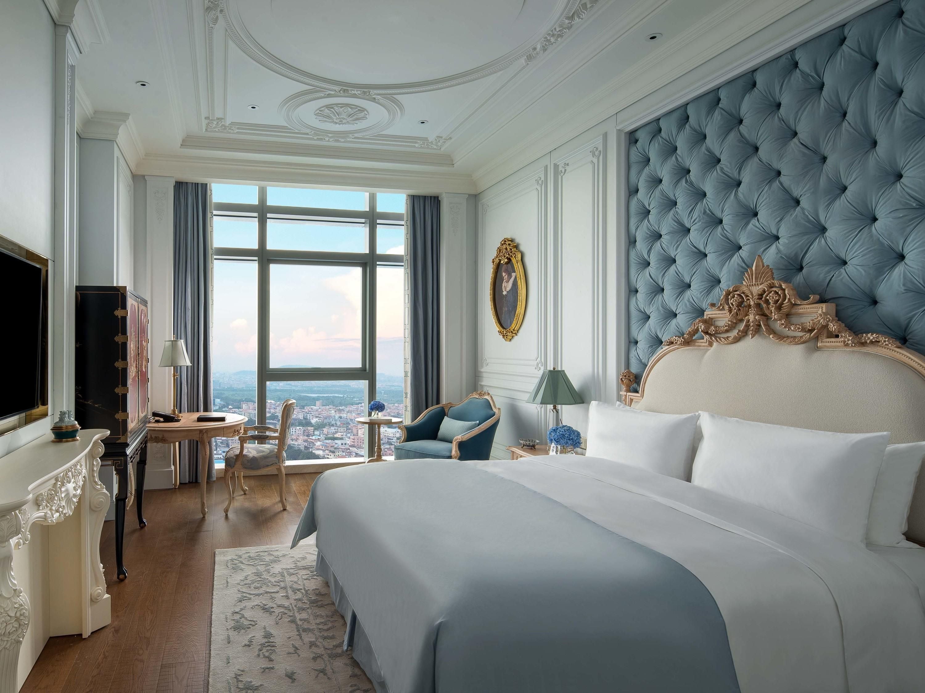 A luxurious hotel room with a large bed, a television, a desk with a lamp, and a view of the city through large windows.