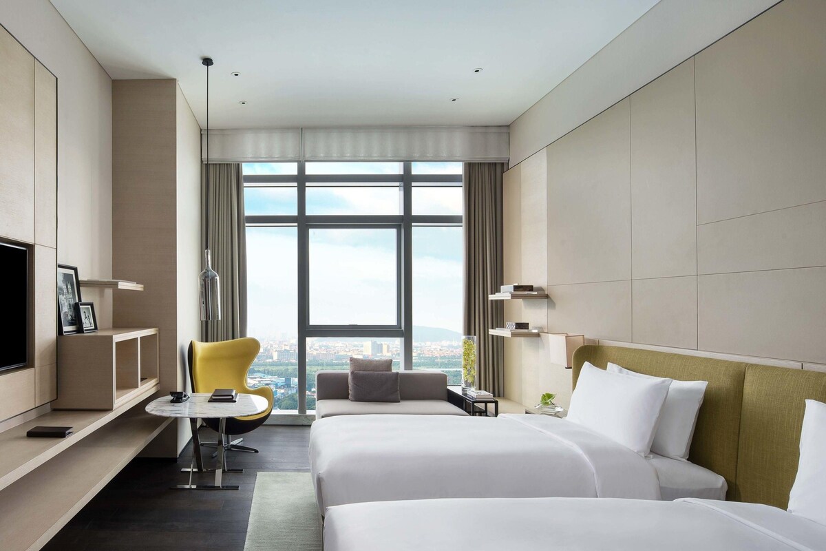 Ein Hotelzimmer mit zwei Betten, einem Fernseher, einem Schreibtisch, einem Sessel und einem großen Fenster mit Blick auf die Stadt.