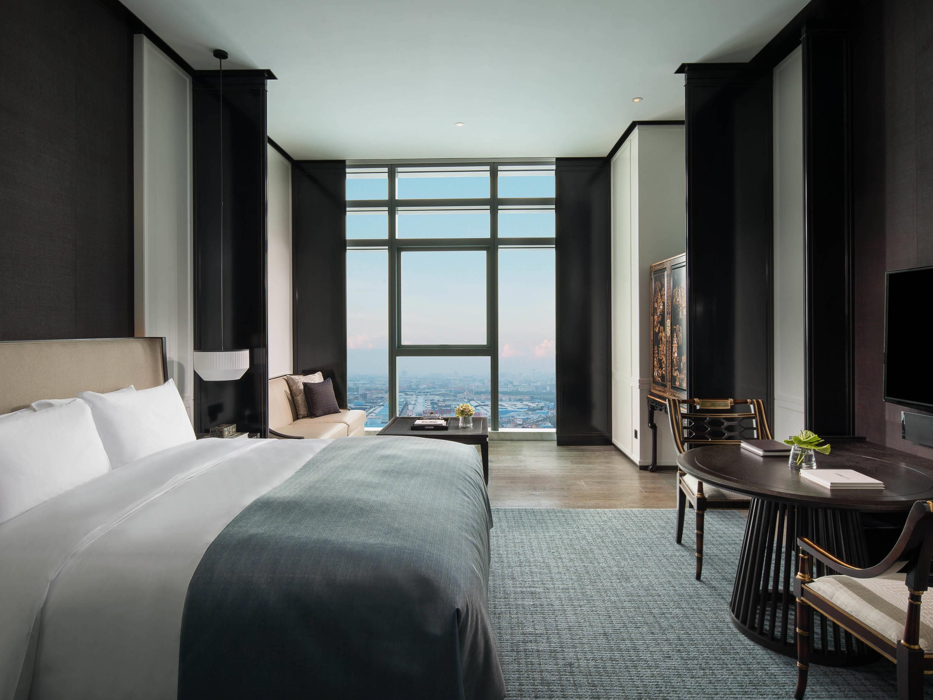 A modern hotel room with a large bed, a desk with a chair, a television, and a view of the city through large windows.