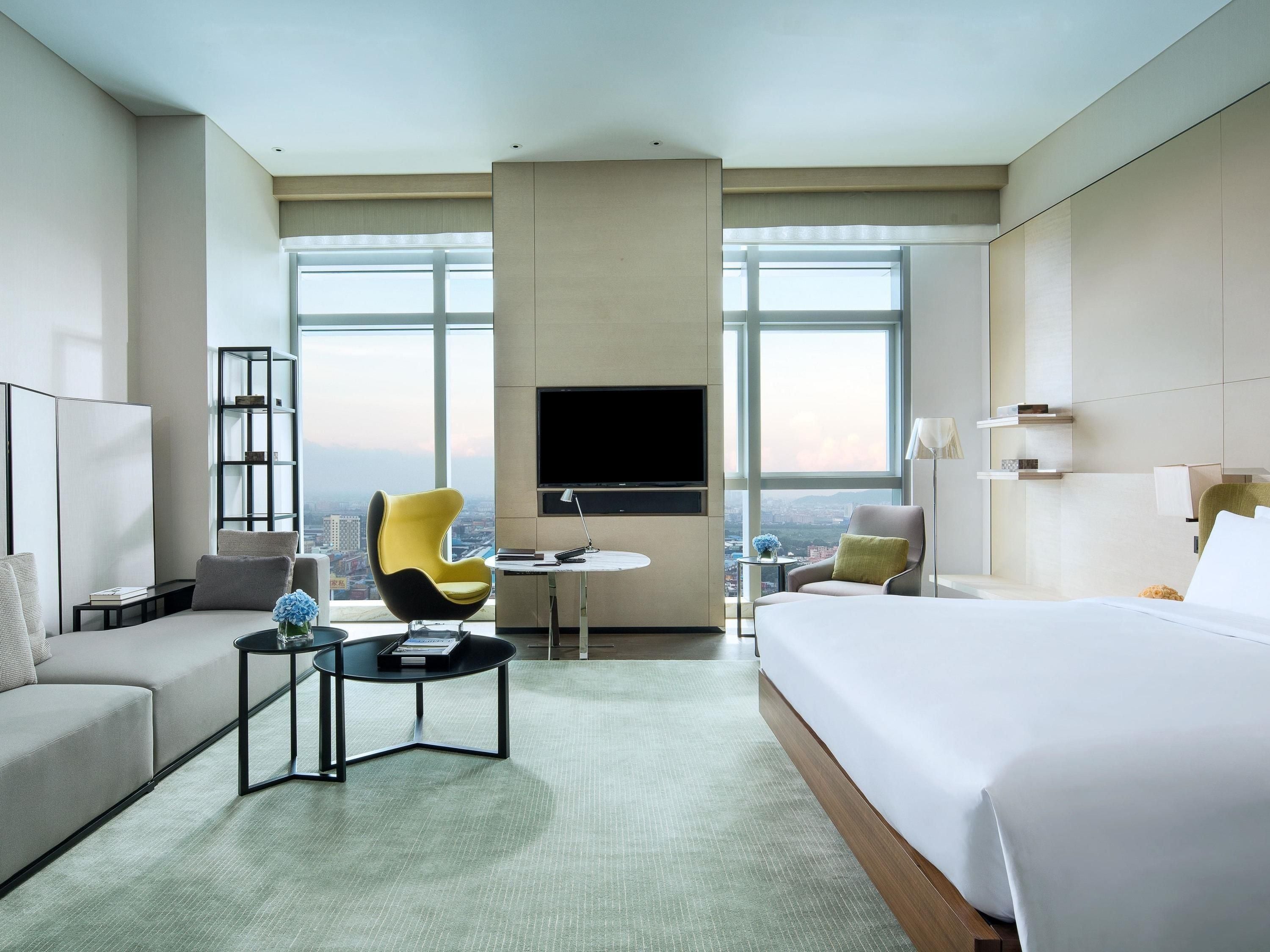 Habitación de hotel con una cama grande, un sofá, una silla, una mesita, un televisor y vistas a la ciudad.