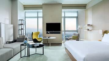 Habitación de hotel con una cama grande, un sofá, una silla, una mesita, un televisor y vistas a la ciudad.