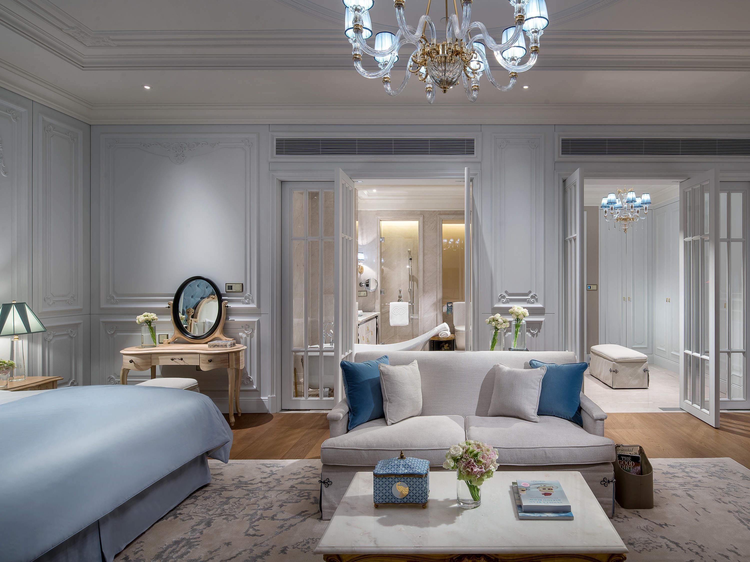 A hotel room with a bed, a sofa, a coffee table, a nightstand, and a mirror. There is a chandelier, a window, and a door leading to the bathroom.