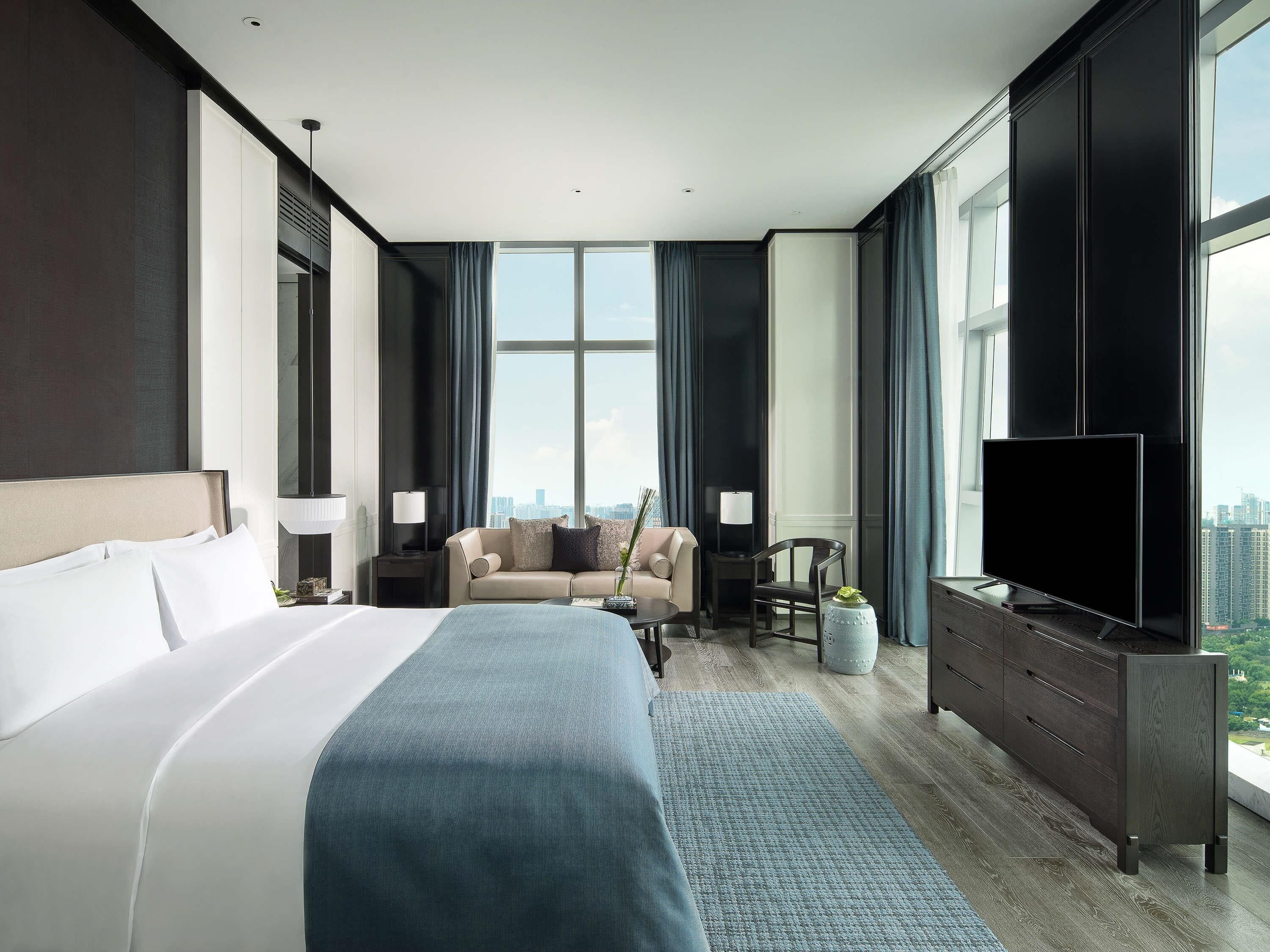 A modern hotel room with a large bed, a sofa, a TV, and a city view.