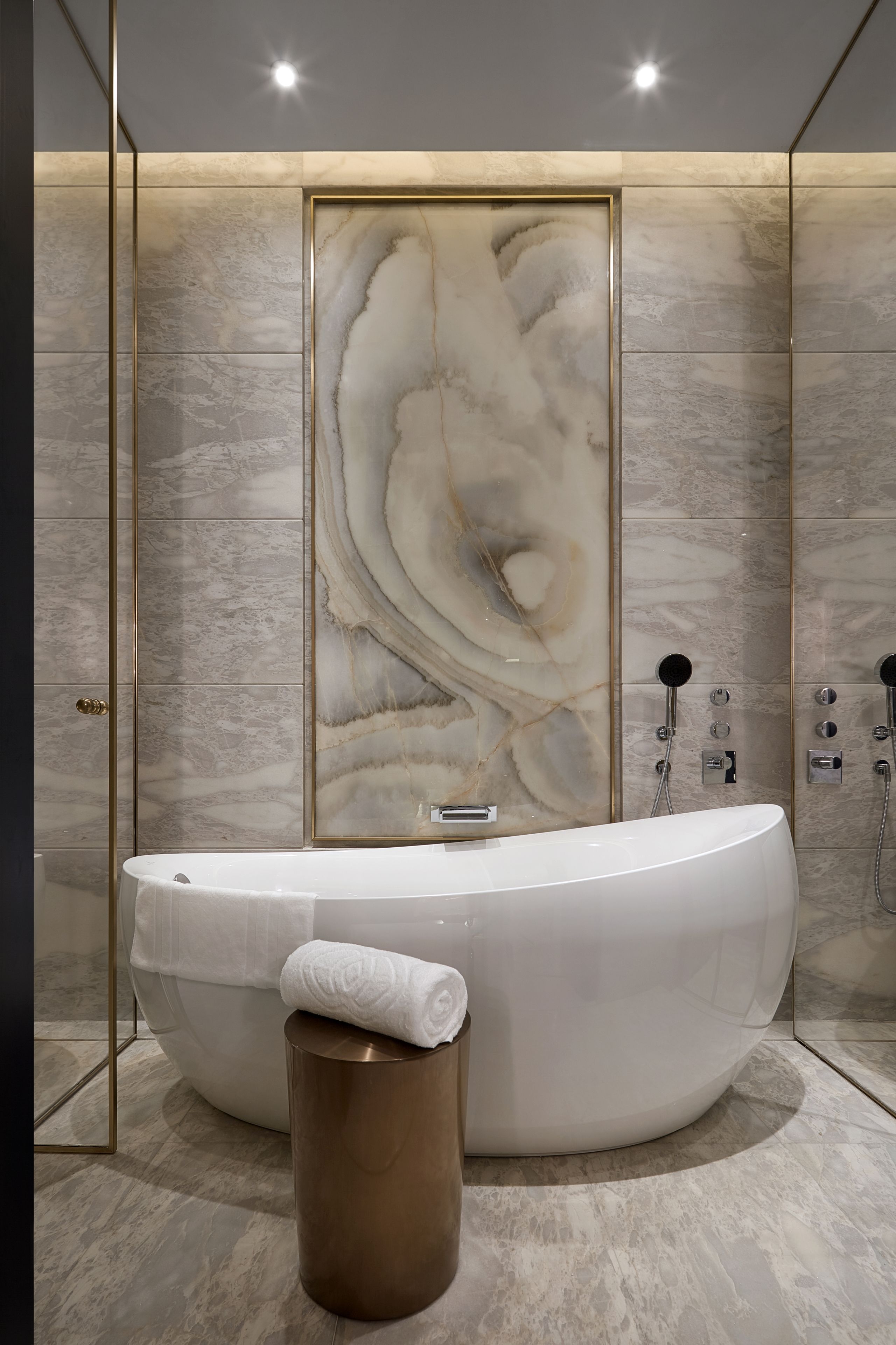 Une salle de bain moderne avec une grande baignoire de forme ovale, un mur en marbre orné d’un motif tourbillonnant et un miroir encadré d’or.