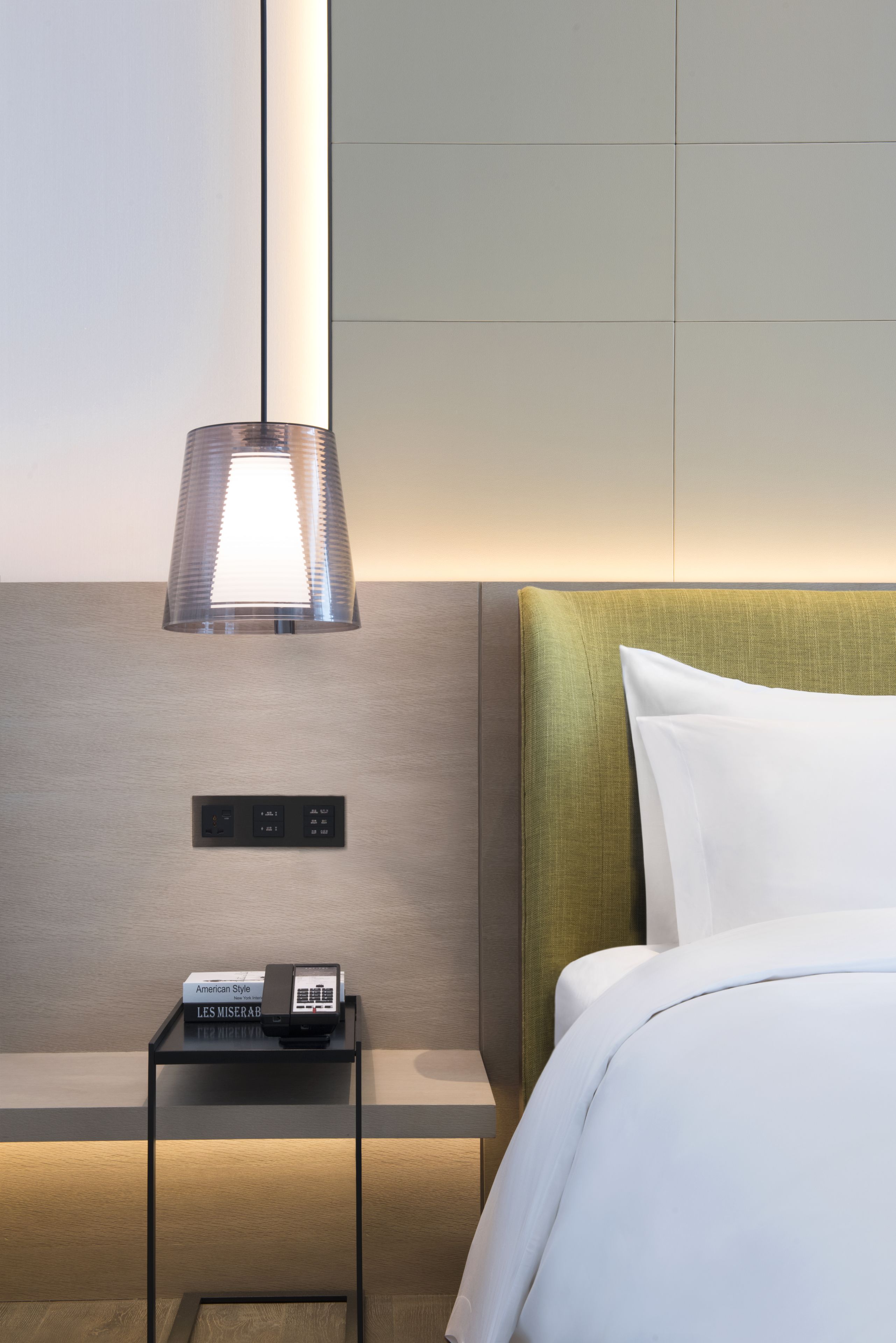 A modern hotel room with a neatly made bed, a bedside table with a lamp, and a control panel on the wall.