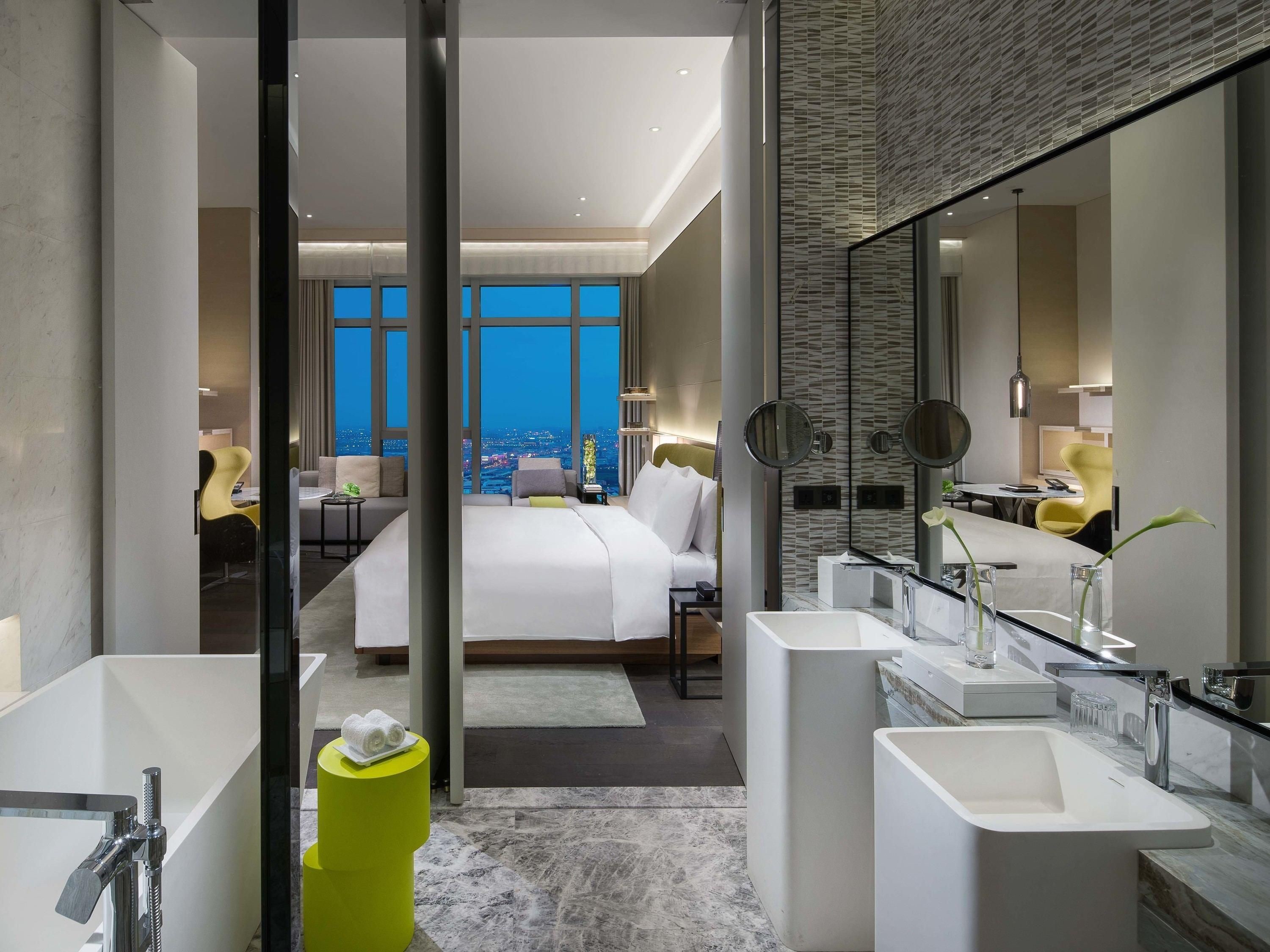 A modern hotel room with a large bed, two side-by-side bathtubs, a large mirror, and a view of the city at night.