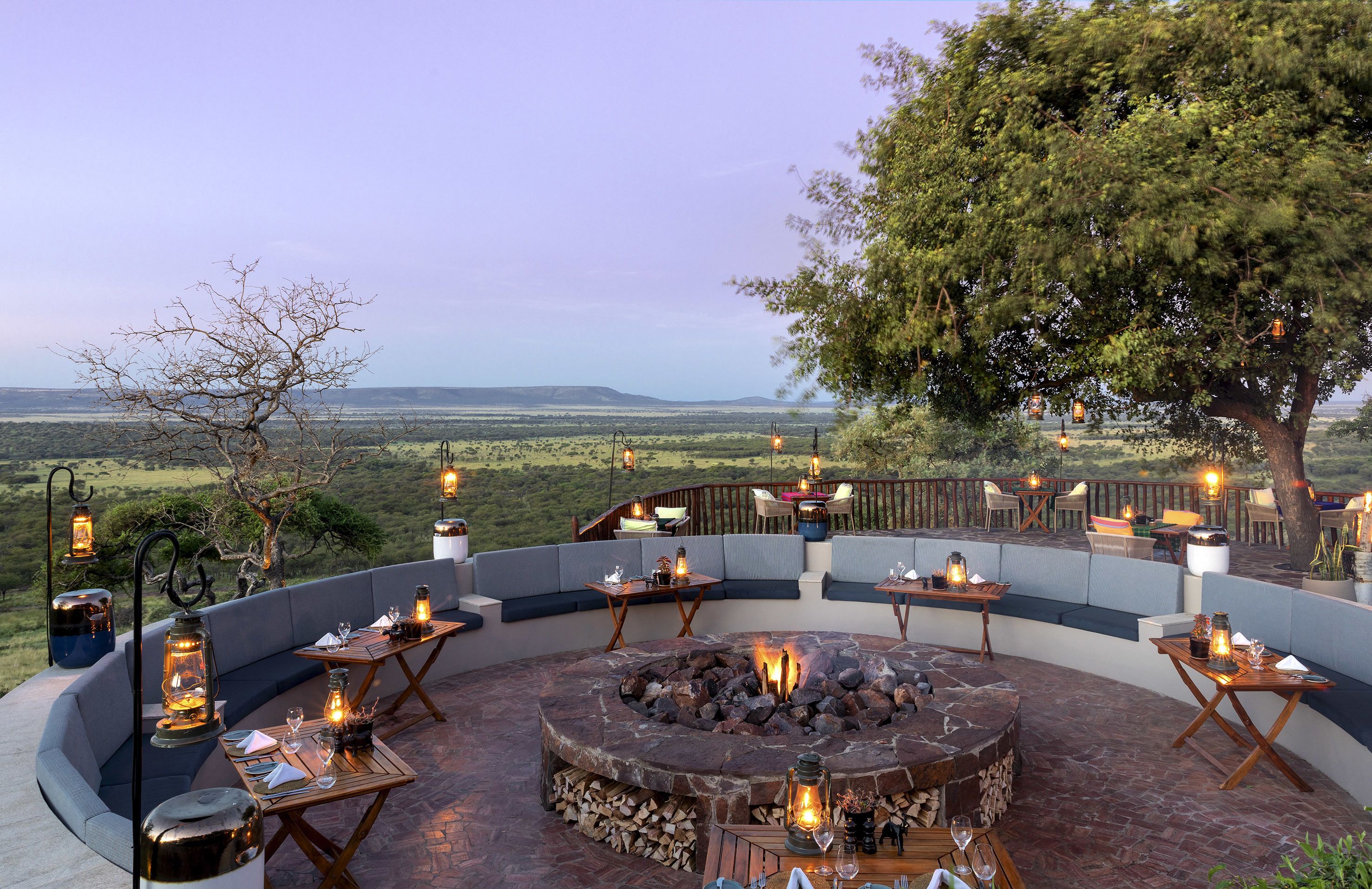 An outdoor seating area with round tables, benches, and lanterns, surrounded by a scenic view.