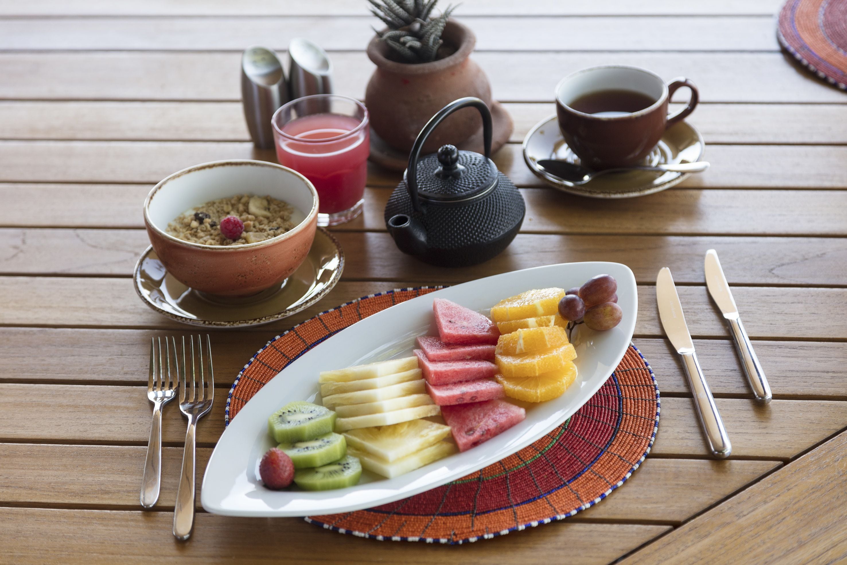 Un desayuno servido en una mesa de madera, con una fuente de fruta, un hervidor de agua, tazas y cubiertos.