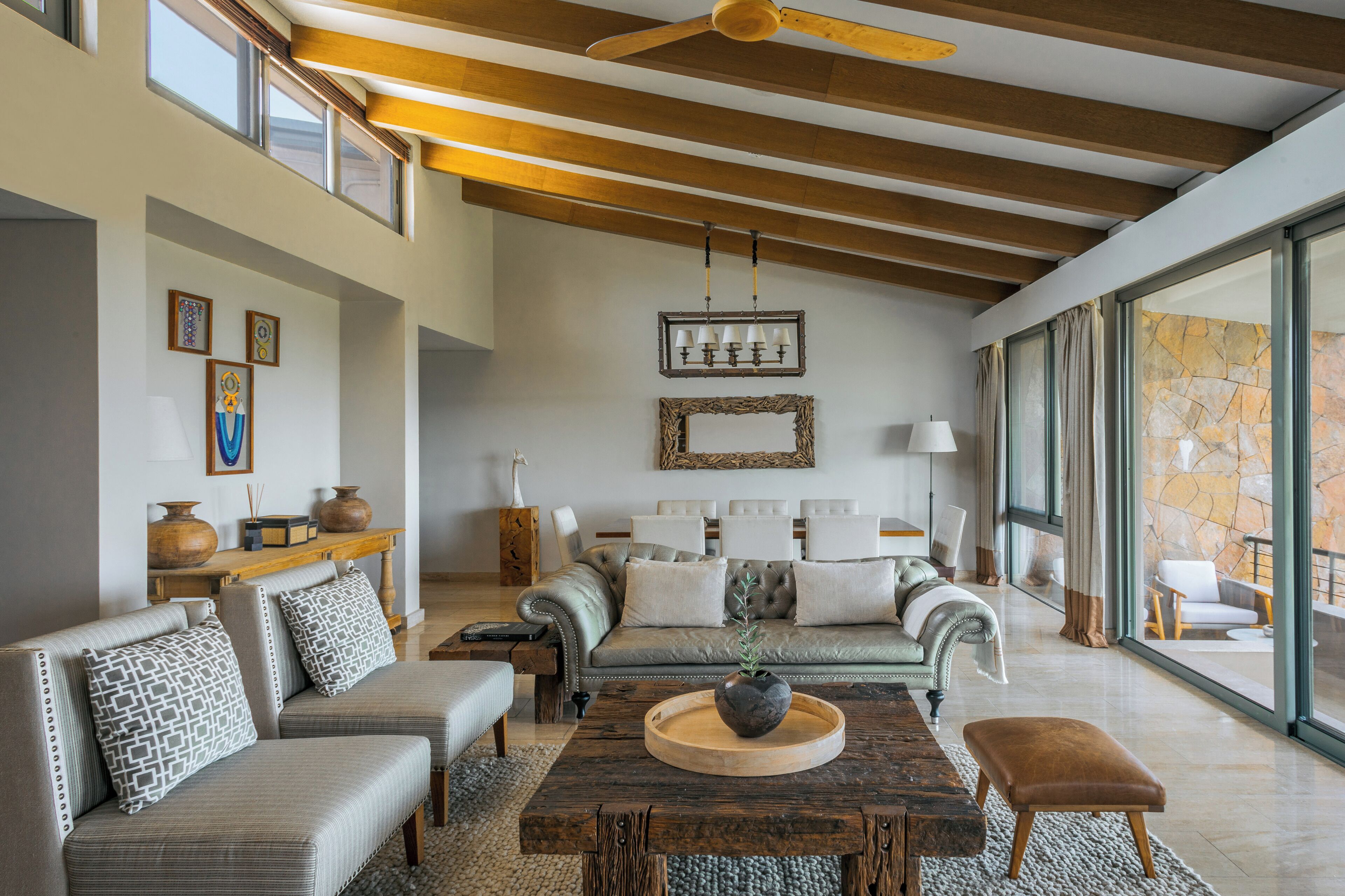 A spacious living room with a high ceiling, wooden beams, and large windows offering a view of the outdoors.