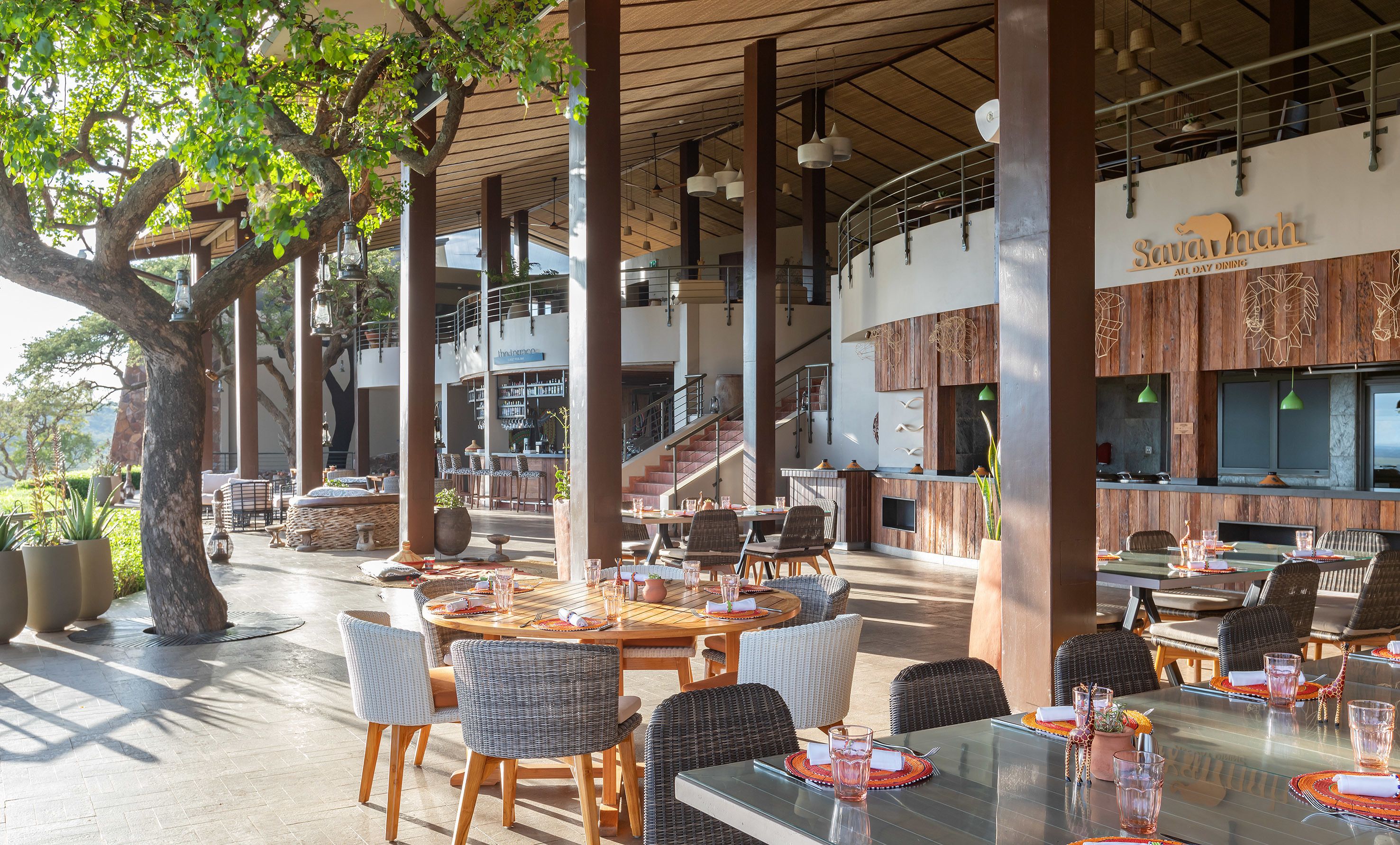 An outdoor dining area with round tables and wicker chairs, a bar area with a sign reading "Savannah All Day Dining", and a staircase leading to an upper level.