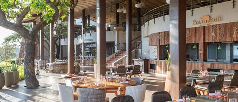 An outdoor dining area with round tables and wicker chairs, a bar area with a sign reading "Savannah All Day Dining", and a staircase leading to an upper level.