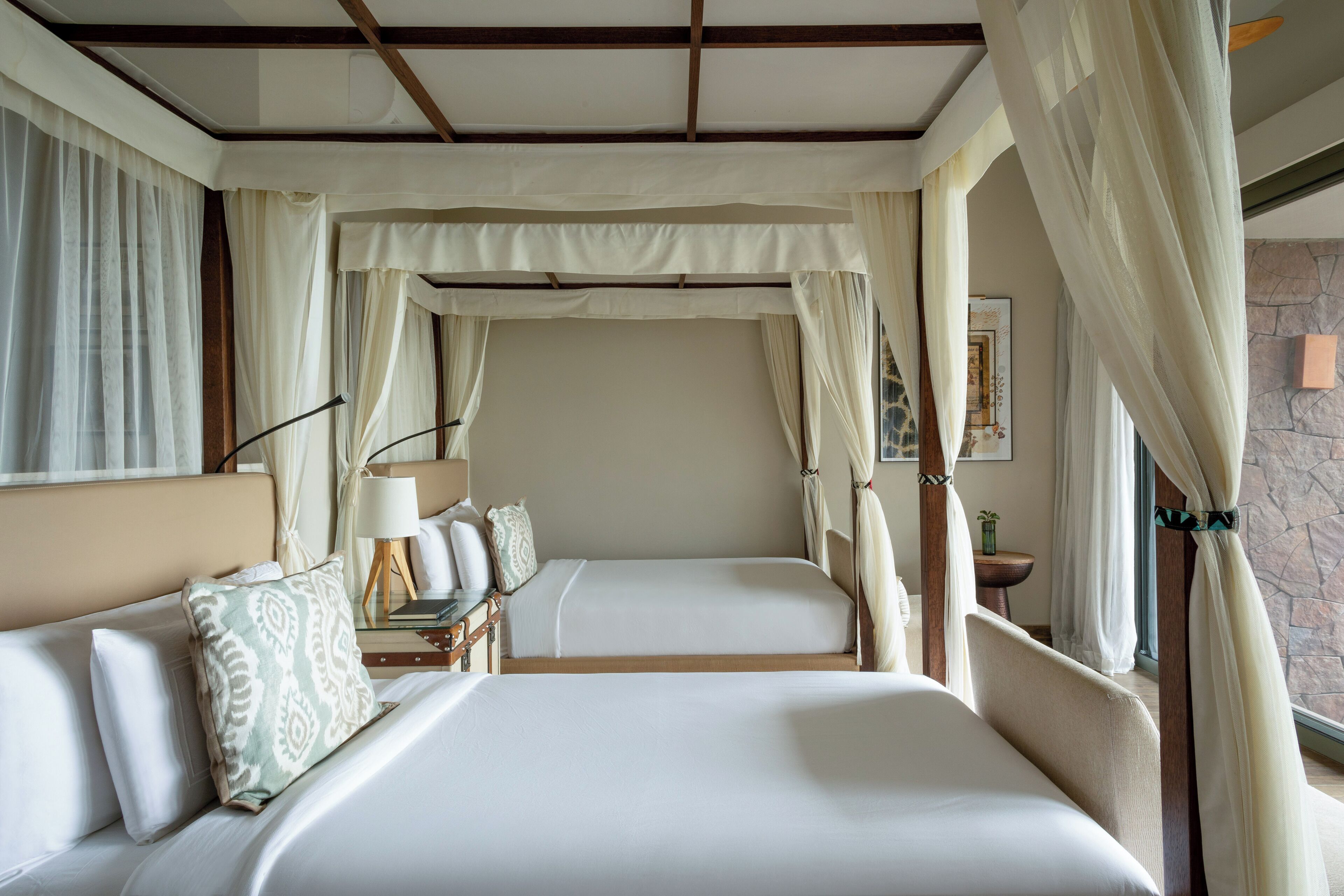 Uma cama de casal com dossel, uma mesa de cabeceira com abajur e vista para uma parede de pedra pela janela.