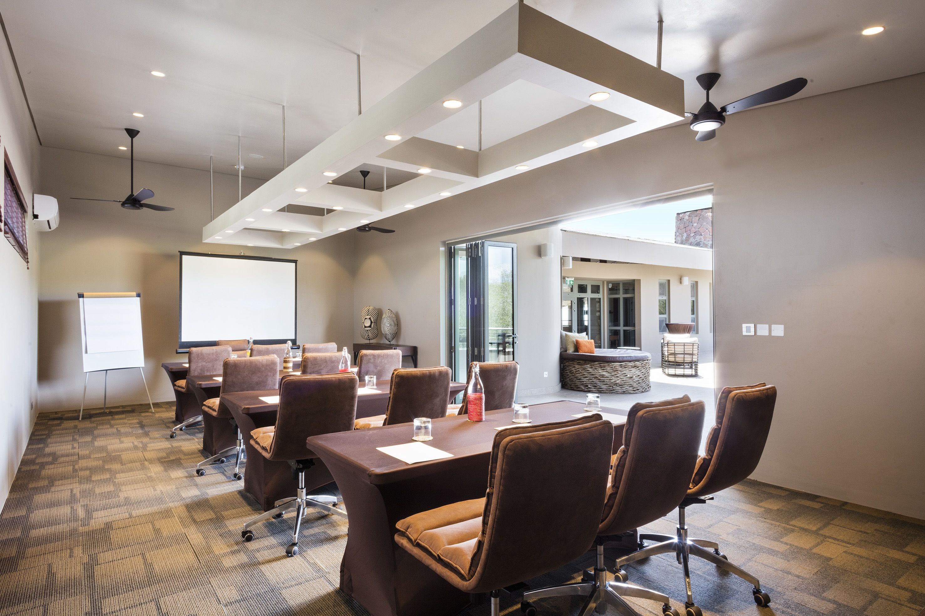 A conference room with a large table, multiple chairs, a projector screen, and ceiling fans.