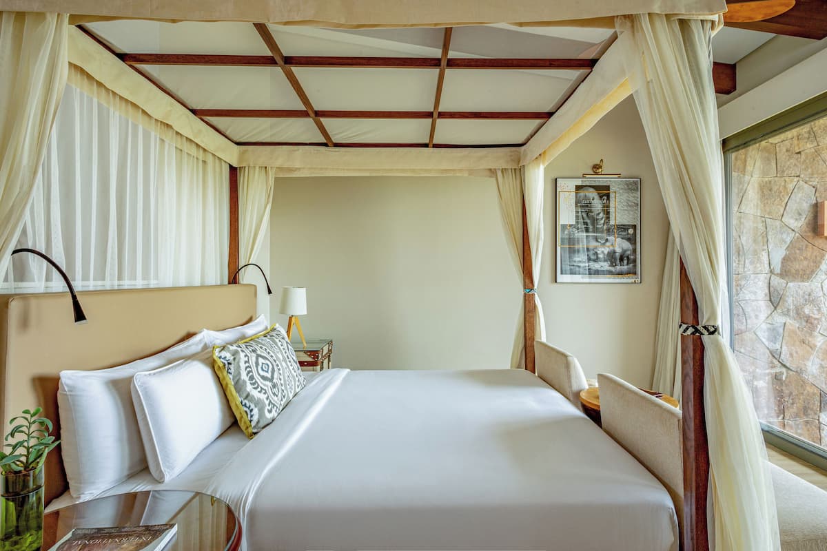 Une chambre d’hôtel avec un grand lit, une table de chevet, une lampe, un tableau encadré au mur et une fenêtre avec des rideaux.