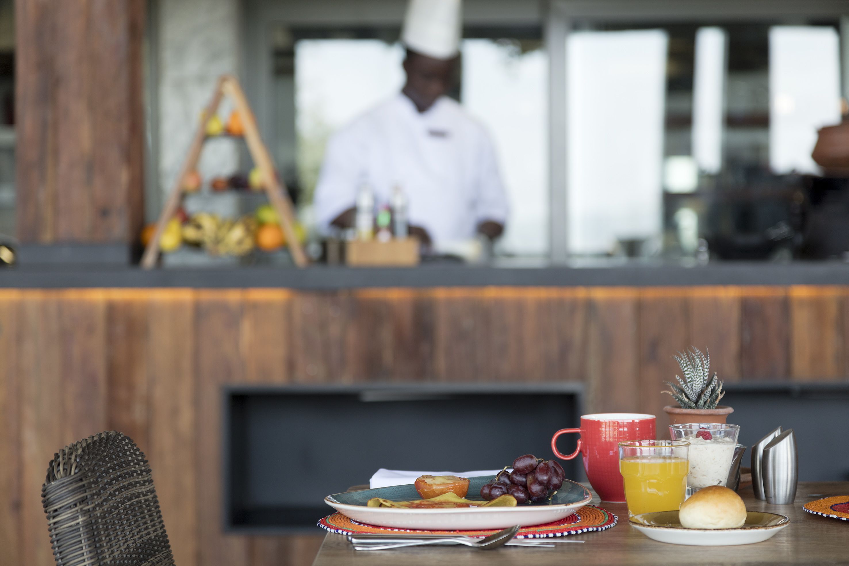A restaurant setting with a wooden counter, a chef in the background, and a table with plates of food, a glass of orange juice, and a small potted plant.