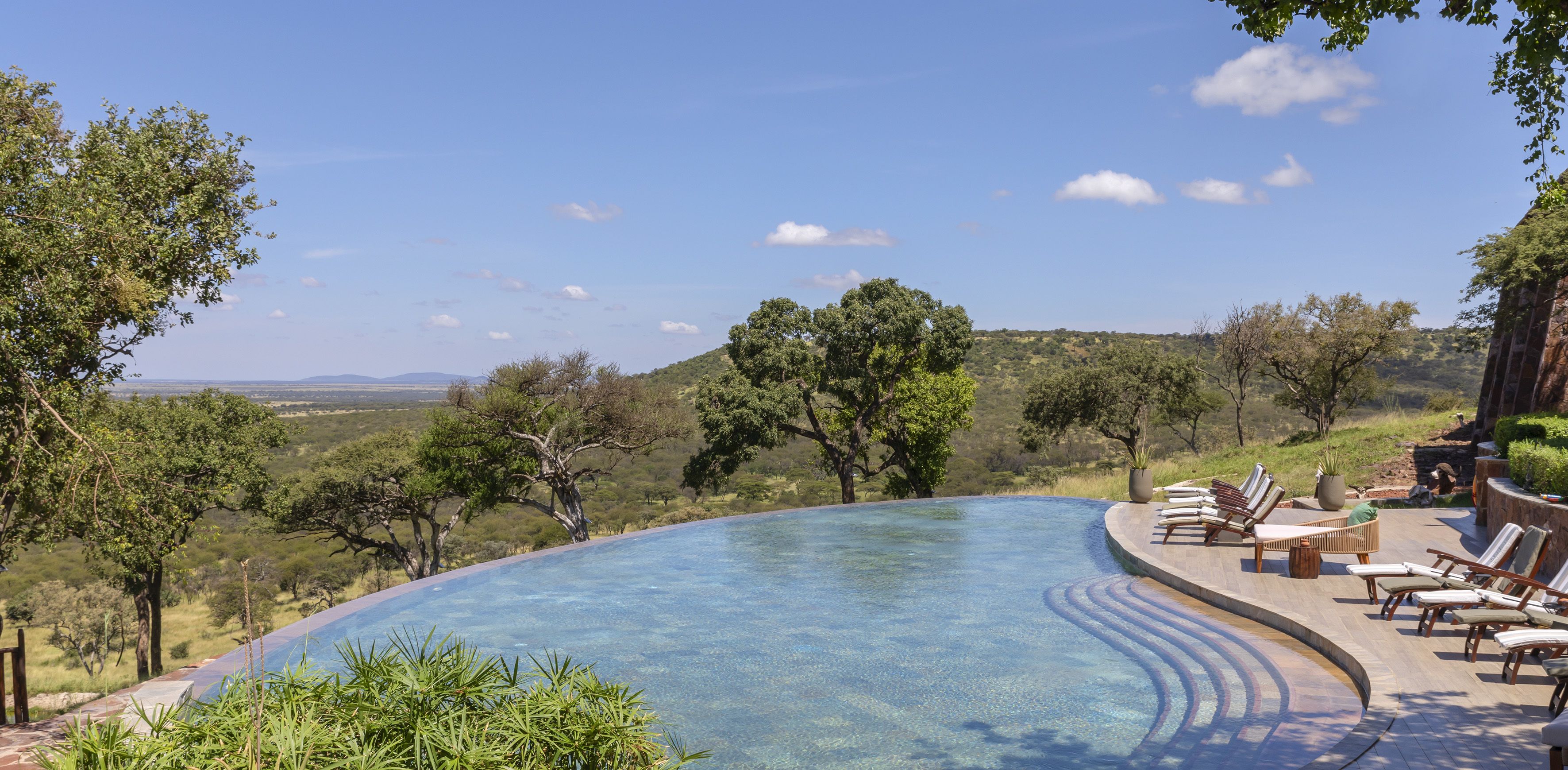 A curved infinity pool with lounge chairs and a view of a hilly landscape.