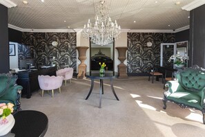 A spacious living room with a chandelier, two green armchairs, a round table with a vase, and a fireplace.
