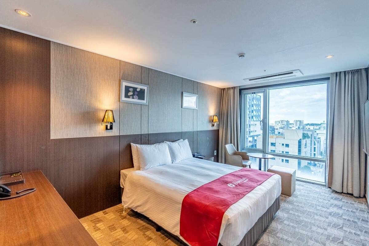 Habitació d'hotel amb un llit gran, un escriptori i vista a la ciutat a través d'un gran finestral.