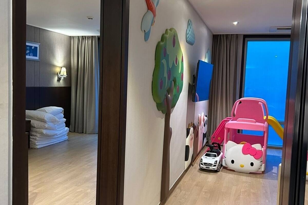 Habitació d'hotel amb sòl de fusta, llit amb roba de llit blanca, escriptori amb làmpada, finestra amb cortines i zona de jocs infantil amb joguines.