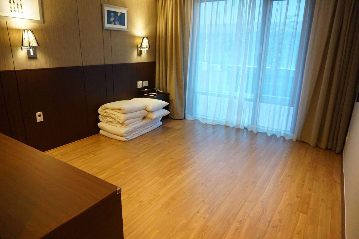 Habitació d'hotel amb sòl de fusta, llit amb tovalloles ordenades, tauleta de nit amb làmpada i finestra gran amb cortines.