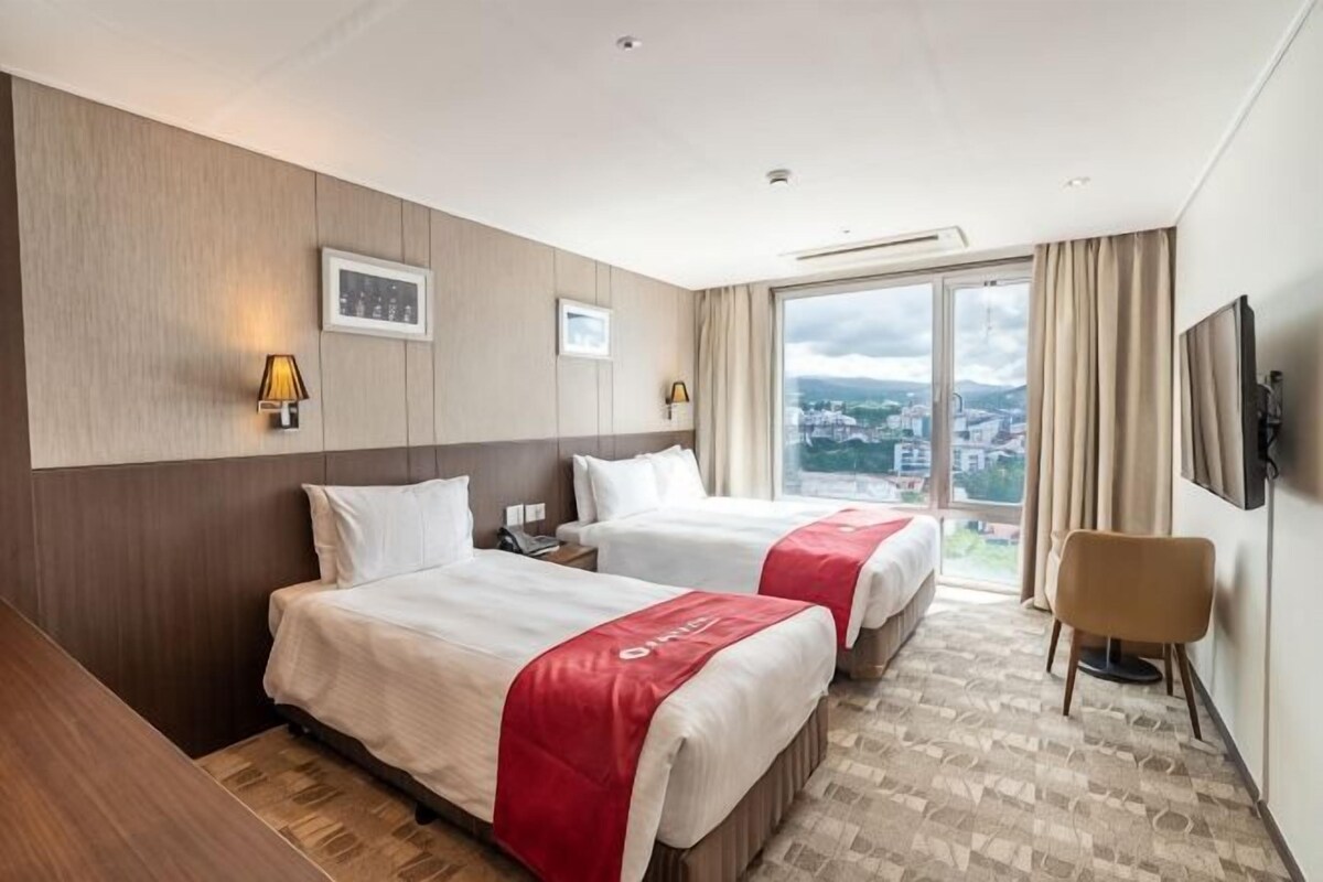 Une chambre d’hôtel avec deux lits, une chaise, un téléviseur et une grande fenêtre donnant sur la ville.