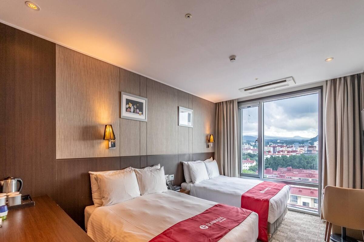 Habitació d'hotel amb dos llits, un panell de capçalera de fusta, una finestra gran amb vista a la ciutat i un llençol vermell sobre un dels llits.