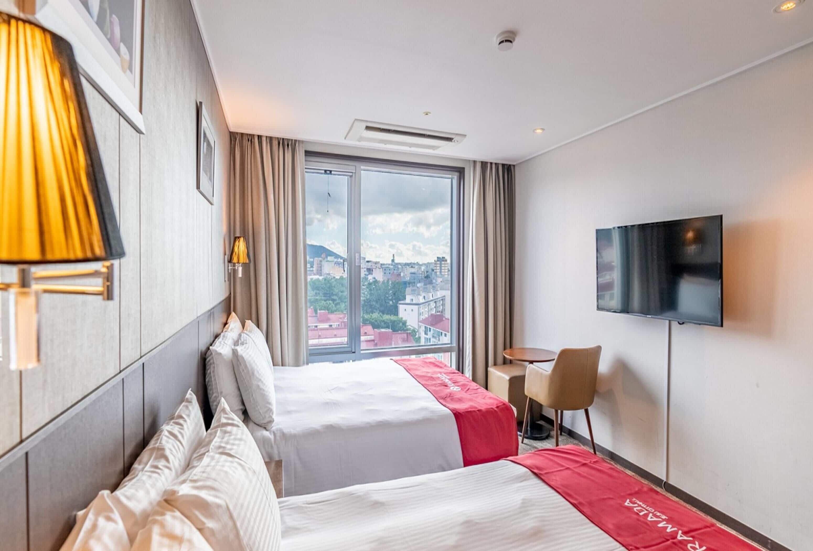 Ein Hotelzimmer mit zwei Betten, einem Fernseher, einem Schreibtisch und Blick auf die Stadt.