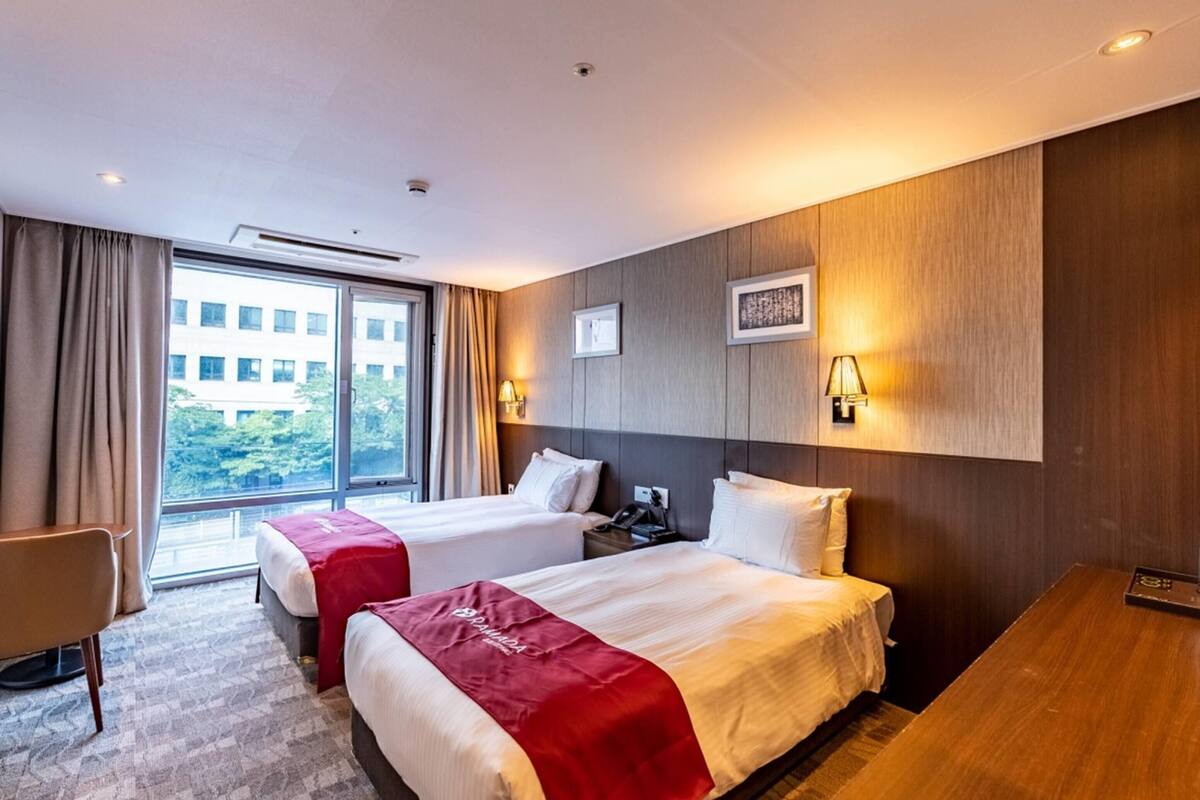 Habitació d'hotel amb dos llits, una taula de treball, una cadira i un gran finestral que ofereix vistes a edificis i vegetació.