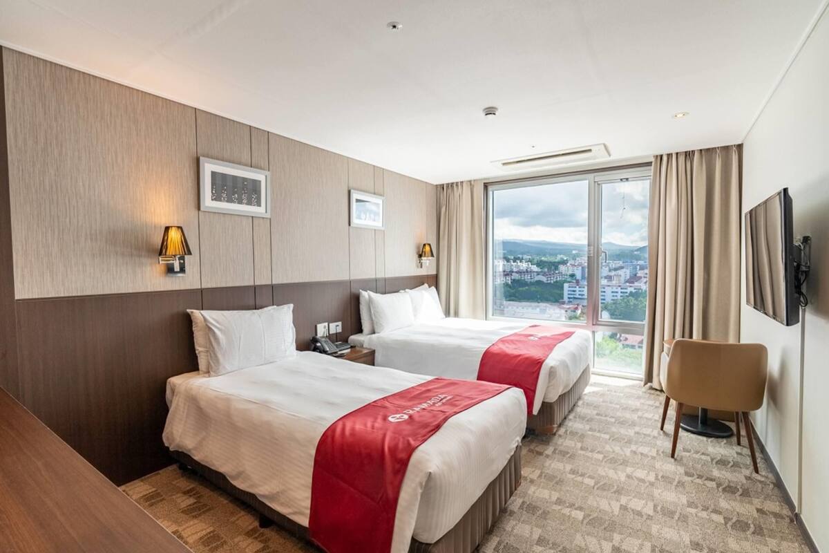 Habitació d'hotel amb dos llits, escriptori, cadira i una finestra gran amb vista a la ciutat.