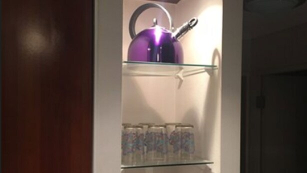 A kitchen with a purple kettle on the top shelf, a glass cabinet below it, and a small table with a box on it. - Departamento Belen Lira (Santiago)