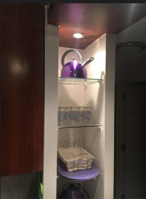 A kitchen with a purple kettle on the top shelf, a glass cabinet below it, and a small table with a box on it. - Departamento Belen Lira (Santiago)