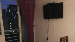 A room with a purple sofa, a small wooden table, a TV mounted on the wall, and a view of a cityscape through a window with a curtain. - Departamento Belen Lira (Santiago)